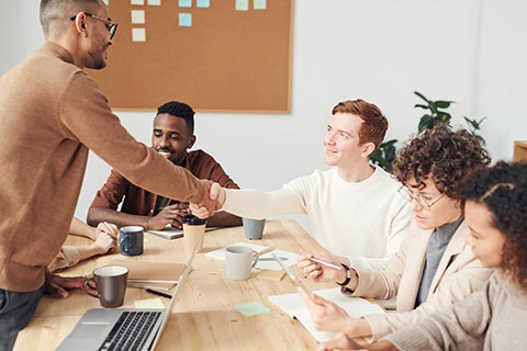A group of colleagues is engaged in a team meeting around a table, with one person standing and greeting others